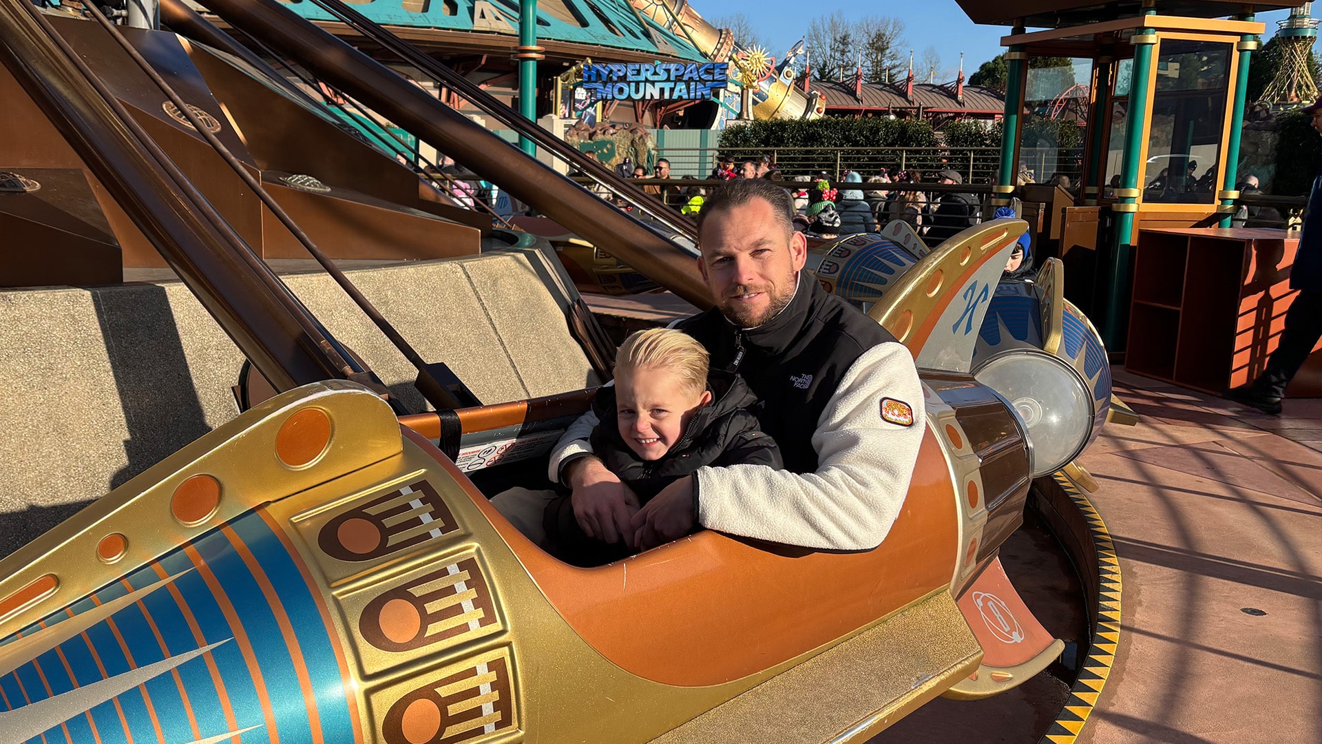 Vader Leon en zoon Jimmy lachen naar de camera vanuit een raket van de attractie Orbitron, met Hyperspace Mountain op de achtergrond.